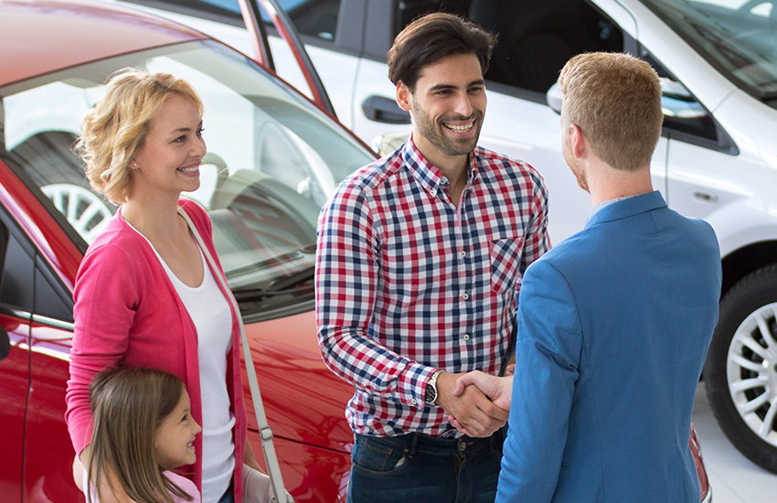 Happy family shaking hands with a car dealership representative in a showroom with a red car in the background.